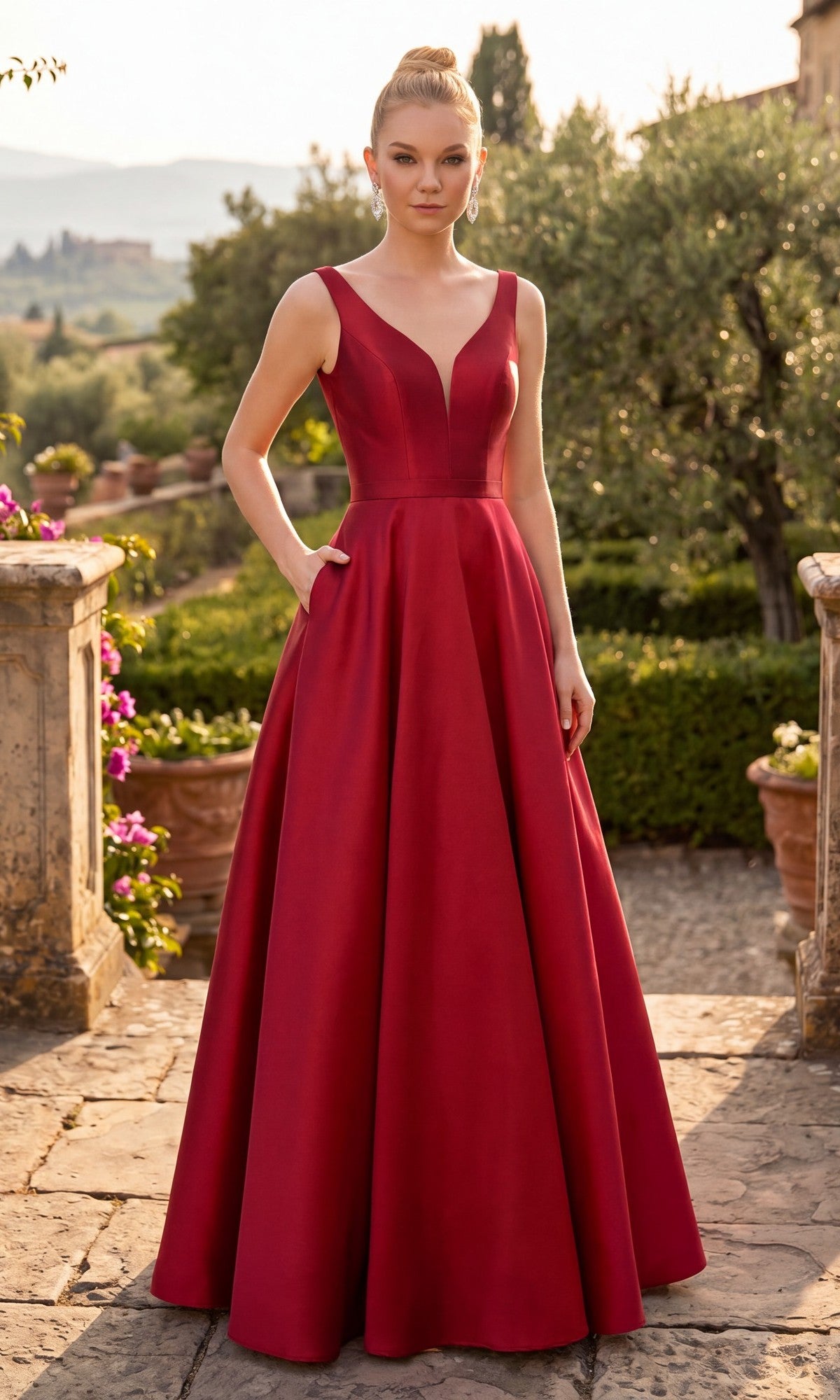 A woman wearing the Long Prom Dress: Chicas C9188 in a rich burgundy shade stands outdoors on a stone path, surrounded by lush greenery and colorful flowers. The elegant gown features a sleeveless fitted bodice with a deep V-neckline and an A-line skirt that flows gracefully to the floor.